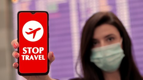 Close up, woman, in protective mask, holds smartphone with airplane icon, stop travel sign, on screen, in front of flight information board in departures terminal, at airport.