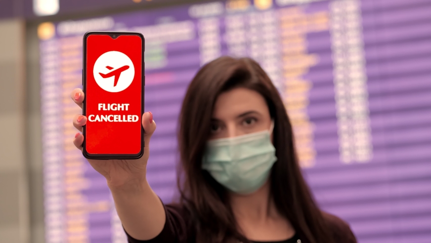Close up, woman, in protective mask, holds smartphone with airplane icon, flight cancelled sign, on screen, in front of flight information board at airport. Royalty-Free Stock Footage #1061747185