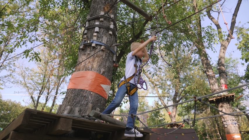 A small six-year-old boy Braves obstacles in a rope Park between trees in the summer.