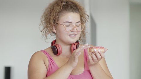 Plus-size coquette woman tasting delicious donut and smiling at camera. Close-up portrait of cheerful Caucasian overweight lady enjoying sweet tasty bakery indoors. Flirt and lifestyle.