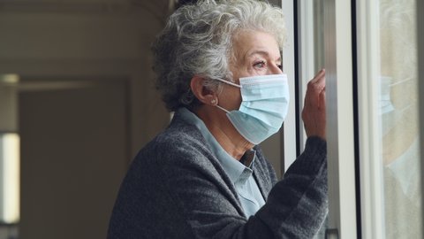 Depressed senior woman with face mask stay at home during Covid-19 pandemic. Old sad woman looking outside of a window with surgical mask during quarantine and lockdown measures for Covid19.