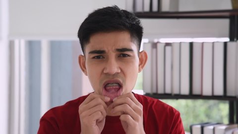 Young Asian man in red casual outfit complains about painful hurt about wearing braces in the living room.