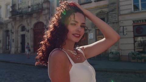 Beautiful curly brunette woman wearing pearl necklaces. Happy, smiling fashionable lady posing in sunny street European city.