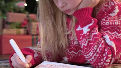 Portrait small blond girl in red sweater lies on floor near Christmas tree and writes letter with markers on paper to Santa Claus. Child writes letter with wishes for gift for Santa Claus