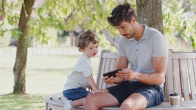 Father sitting on park bench under tree with son looking at video game on mobile phone together - shot in slow motion