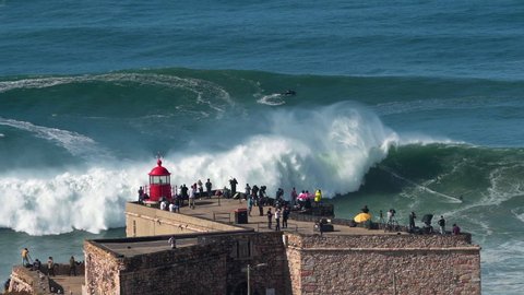 Nazare, Portugal - October 29, 2020: Surfer riding giant wave near the Fort of Nazare Lighthouse in Nazare, Portugal. Nazare is famously known to surfers for having the largest waves in the world.