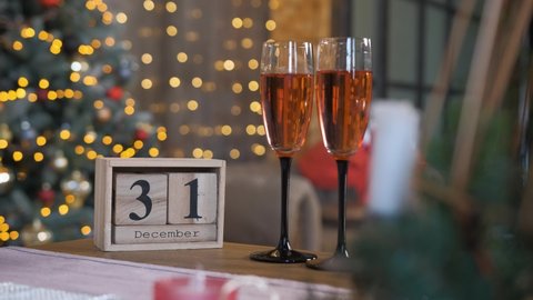 Wooden block calendar and two glasses of champagne on the table, on the background Christmas decoration and beautifully lit Christmas tree. Holiday countdown