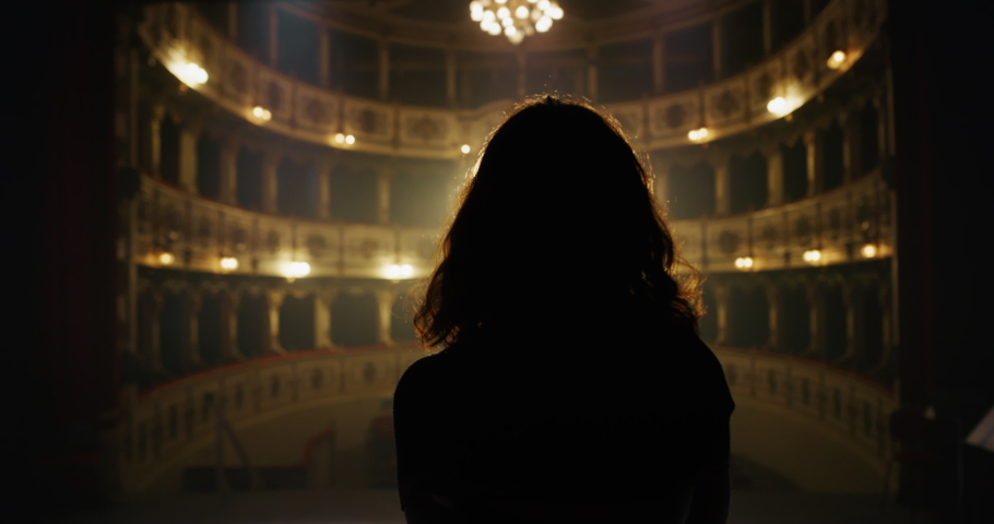 Cinematic Back View of Female Singer or Actress Going on Stage Under a Spotlight in an Empty Classic Theatre. Female Artist Rehearsing for her Big Show Alone. Silhouette Slow Motion Shot Royalty-Free Stock Footage #1061779087