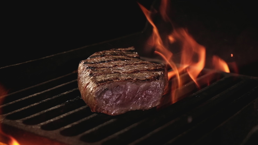 Juicy beef steak is fried on fire coals on iron grill on dark background in flame of smoke. Barbecue in evening, chef turns meat on grill with tongs. cooking dinner in a hot flame. | Shutterstock HD Video #1061784094