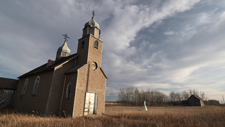 Looking up an old abandoned modest church with faux brick siding and white highlights.  Two orthodox crosses are on top of the church.  Clouds move through a blue sky.
