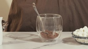 Pouring hot milk in a glass cup, woman prepares hot chocolate with marshmallow, close up video, marble background