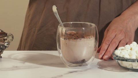 Pouring hot milk in a glass cup, woman prepares hot chocolate with marshmallow, close up video, marble background