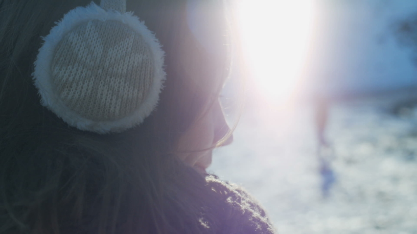 Young woman in fur headphones, sunglasses and turquoise coat, scarf and with backpack enjoys the sun and weather. Around the ski winter resort, snow, mountains, lifts. Slow motion.