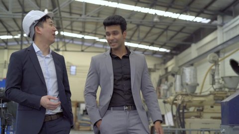 Two young adult diverse businessmen discussing while walking in industrial factory background. Asian and Indian ethnic men in business formal suit talking - Front View - 4K Real Time Footage