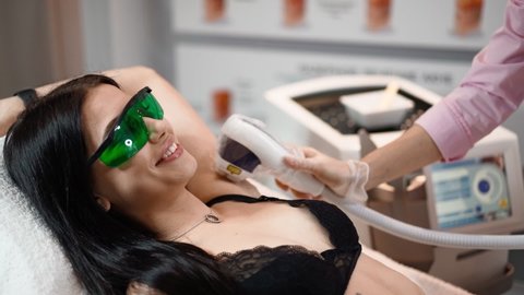 Female beautician doing armpit laser hair removal of attractive sexy woman lying on medical couch and smiles in beauty salon. Cosmitology, epilation and spa concept. Body care. Slow motion. Close-up