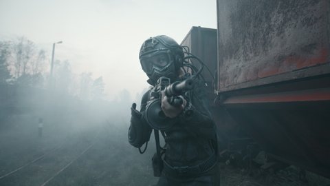 Female soldier of future aims with assault gun and moves towards target by rusty train carriage. Lonely cyborg woman in futuristic combat suit goes through cloud of smoke during battle