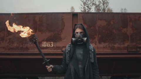 Woman in cyberpunk suit and gas mask standing by rusty train carriage hold burning torch in hand and looking into camera. Dystopian warrior concept, female survivor in abandoned city. Post apocalypse
