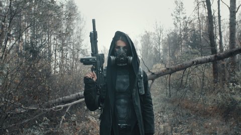 Human in tragic future. Beautiful woman in cyberpunk clothing and grunge gas mask standing in dead forest. Dystopian stalker concept, female survivor posing with assault gun. Post apocalyptic world