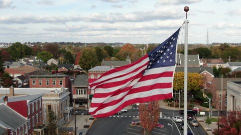 Manheim , PA / United States - 10 18 2020: Close up of American Flag flying above small town in USA. Businesses line town square while traffic drives by. Aerial drone shot.