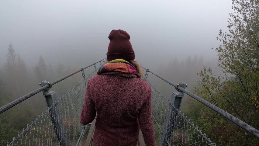 A young woman crossing a bridge above a small, foggy valley
