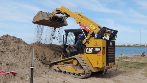 PORT ARANSAS, TX - 29 FEB 2020: Working man drives a CAT 289D front loader to empty a load of dirt at a construction site near the water.