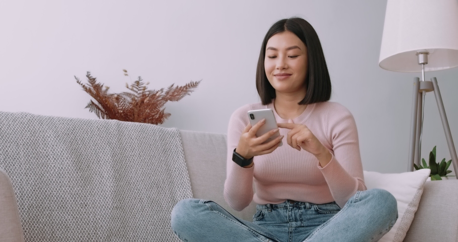 Young asian lady surfing social media on mobile phone, enjoying amazing news , sitting on sofa at home | Shutterstock HD Video #1061809531