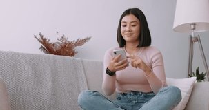 Young asian lady surfing social media on mobile phone, enjoying amazing news , sitting on sofa at home