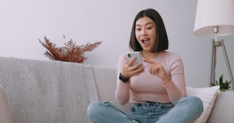 Young asian lady surfing social media on mobile phone, enjoying amazing news , sitting on sofa at home | Shutterstock HD Video #1061809531