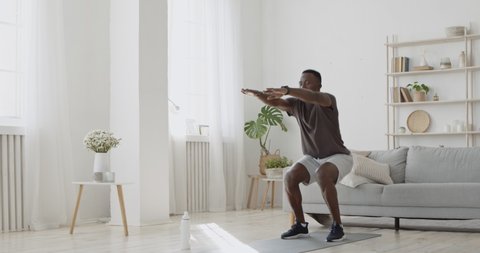 Home workout. Young sporty african american man doing squats exercises in living room interior, sports training alone