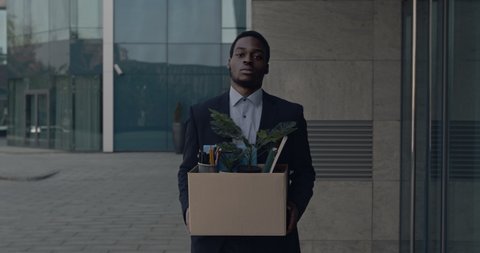 Depressed african american male office worker leaving business center building with box of personal stuff. Businessman lost job. Fired man walking outdoor. Depressed jobless person. Unemployment.