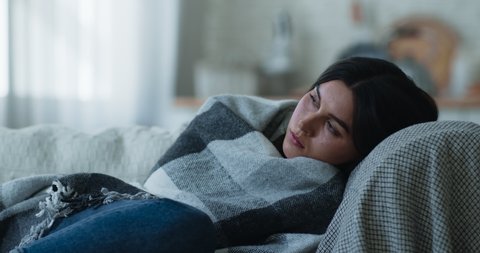 Sad depressed young woman lying alone on sofa wrapped in plaid, feeling tired lonely vulnerable and weak, thinking of problem, looking at camera