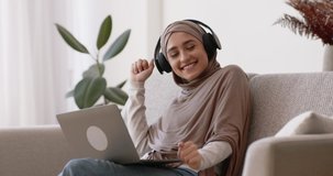 Overjoyed muslim woman in hijab listening music in wireless headphones and dancing, networking on laptop on couch at home