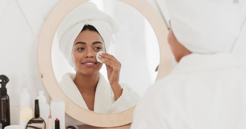 Focused young african american woman applying toner, feeling excited with skin condition Black lady using cotton disposable pads, moisturizing skin before starting makeup.