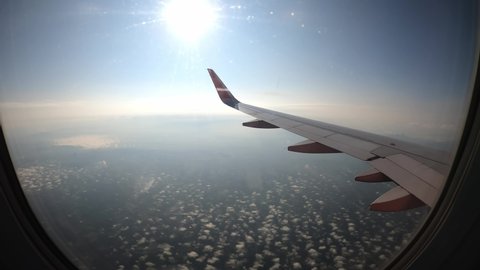 Time lapse of aerial view from airplane window seat, clear blue sky with shining sun rays, airplane wing moving above puffy white clouds and city landscape. Airplane smooth landing on airport runways.
