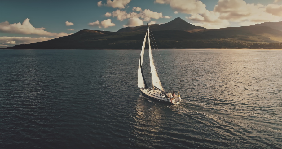 Aerial of sun yacht sail in ocean bay. White boat at open sea. Summer cruise on sailboat at sunlight with clouds. Scotland island of Arran sea coast with water transport. Serene seascape Royalty-Free Stock Footage #1061813485