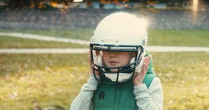 Boy puts helmet on his head. Cheerful happy child playing American football outdoors in sunny day at public park. Family sports weekend. 4K video.