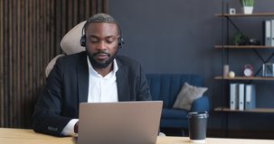 Handsome African American Operator in Call-Center, Technical Support is working Indoor, sitting at Home Office. Handsome friendly Man is talking speaking in Headset, using Laptop. Online Job.