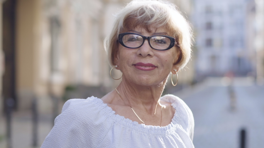 Close-up portrait of smiling confident senior woman in eyeglasses posing outdoors in sunlight. Happy Caucasian female retiree enjoying pension on city street. Leisure and happiness. | Shutterstock HD Video #1061818351