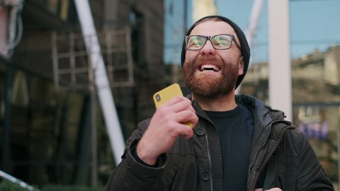 Crop view of man in glasses with bag on shoulder looking at phone screen while walking at street. Bearded male person using smartphone and rejoicing while having good news