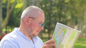 Video, an emotional adult man in sunglasses looks at a city map and looks for direction on the street on a sunny day.