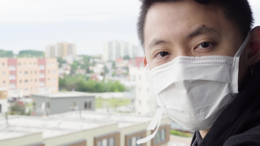 A young Asian man in a face mask looks at the camera as he stands on a balcony in an urban area - face closeup | Shutterstock HD Video #1061821285