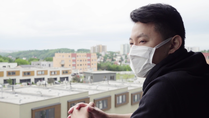 A young Asian man in a face mask stands on a balcony and looks at a cityscape below, thoughtful - closeup | Shutterstock HD Video #1061821300