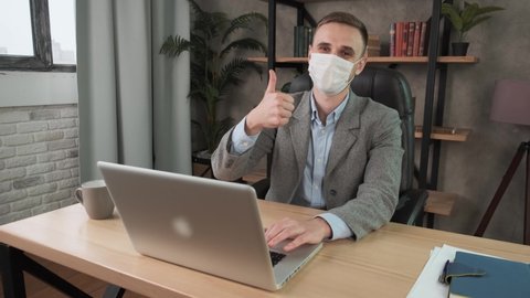 Young confident man employee in a medical facial mask works in the office at the computer. Male worker looking at camera and showing thumb up, working remotely at home. Work during self-isolation and