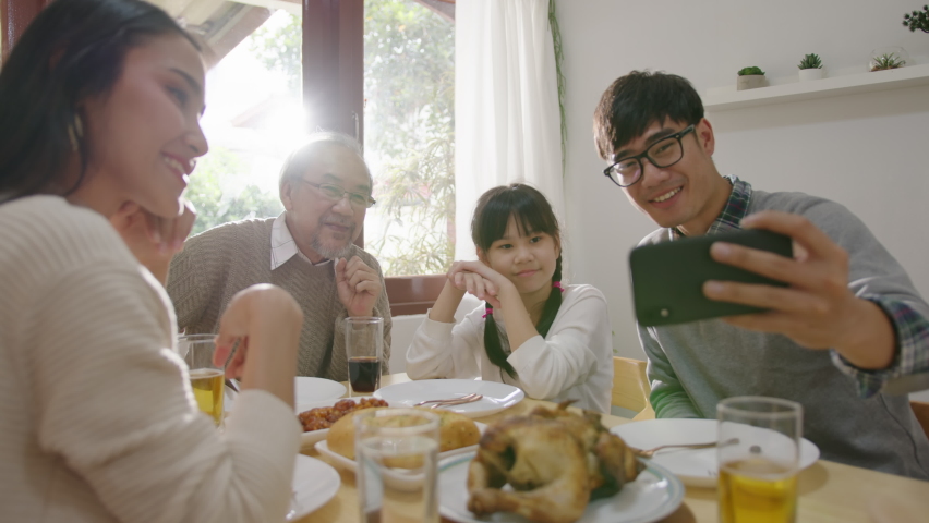 Candid of happy asian family having fun on dining table at home holding mobile video call online and selfie or take photo shoot group with smile and laugh together. Royalty-Free Stock Footage #1061823043