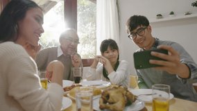 Candid of happy asian family having fun on dining table at home holding mobile video call online and selfie or take photo shoot group with smile and laugh together.