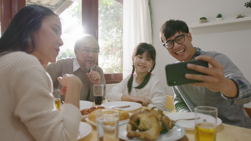 Candid of happy asian family having fun on dining table at home holding mobile video call online and selfie or take photo shoot group with smile and laugh together. | Shutterstock HD Video #1061823043