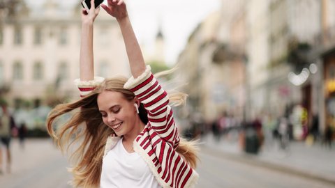 Young adult happy woman dancing in downtown very emotional. Slow motion of happy young woman in wireless headphones dancing singing outdoors in city street having fun alone.