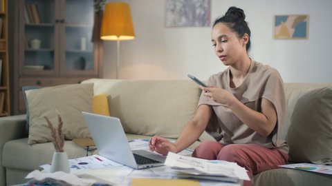 Medium shot of young attractive businesswoman talking on phone while doing paperwork opening envelope with printed diagrams and charts about finance accounting entering data in laptop