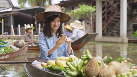 Asian market woman using smartphone to sell food online on website with technology internet media in floating market on boat near canal. Modern and old lifestyle, Ratchaburi District, Thailand.