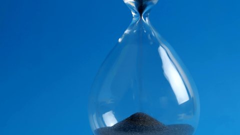 Extreme close-up of black sand running down through hourglass on a blue background. A concept the sand runs out to the hourglass representing run out of time. 4k